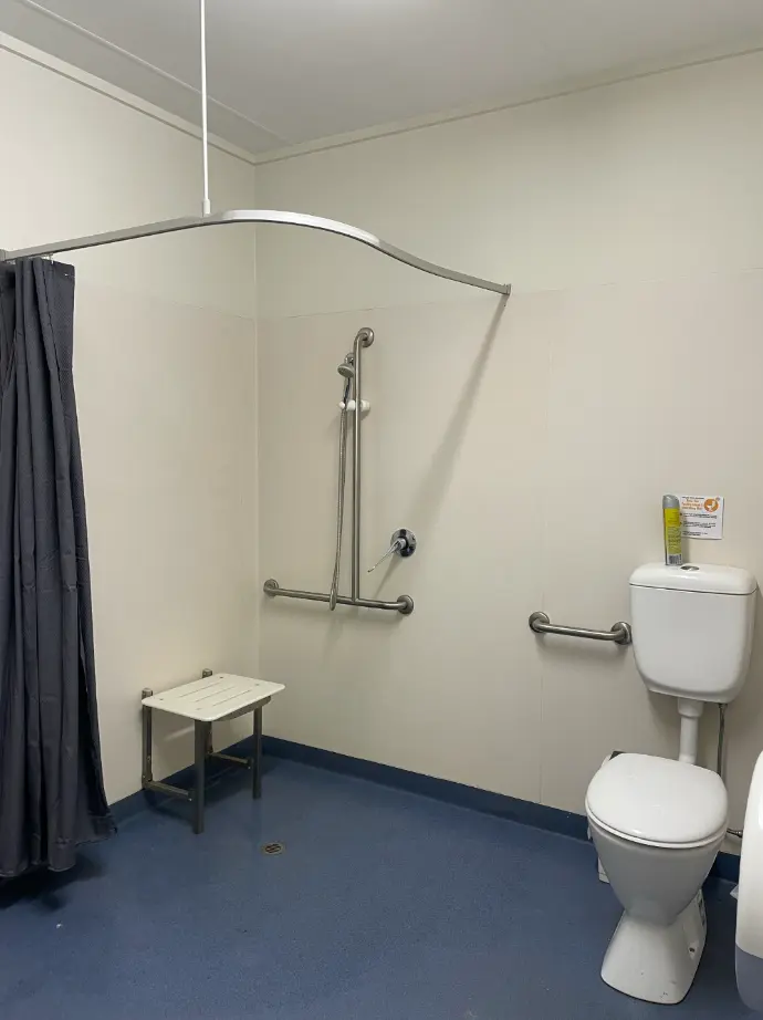 Picture of the Accessible Bathroom from further inside - Showing the roll in shower with open shower curtain, seat folded down, shower head on flexible hose, and a toilet with handrails along the back wall in the shower, and between the shower and toilet.