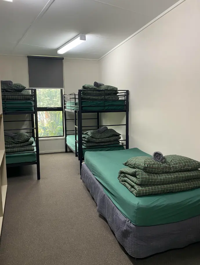 Image shows room layout with two bunk beds and a king single, with folded linen sitting on top of each bed. A window is centred on the far wall, with the blind 30% drawn. The mattresses are all covered with dark green fitted sheets.